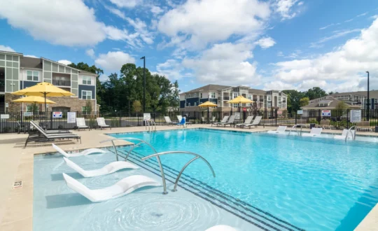 Outdoor swimming pool with lounge chairs and umbrellas, surrounded by apartment buildings under a blue sky.