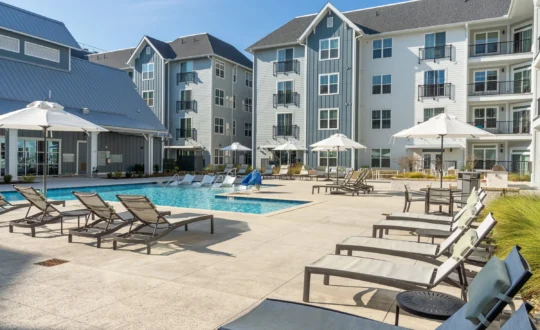 Outdoor apartment pool area with lounge chairs, umbrellas, and modern multi-story buildings in the background.