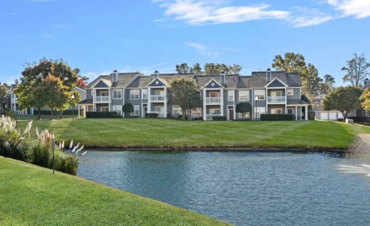 Row of apartment buildings with balconies, seen across a pond and green lawn on a sunny day.